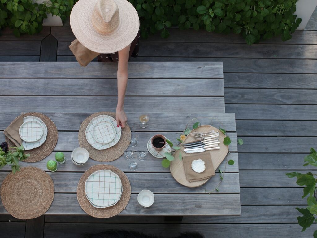 En person dukar ett utomhusbord med tallrikar, glas och servetter. Borden har naturliga linser och gröna växter runtom.