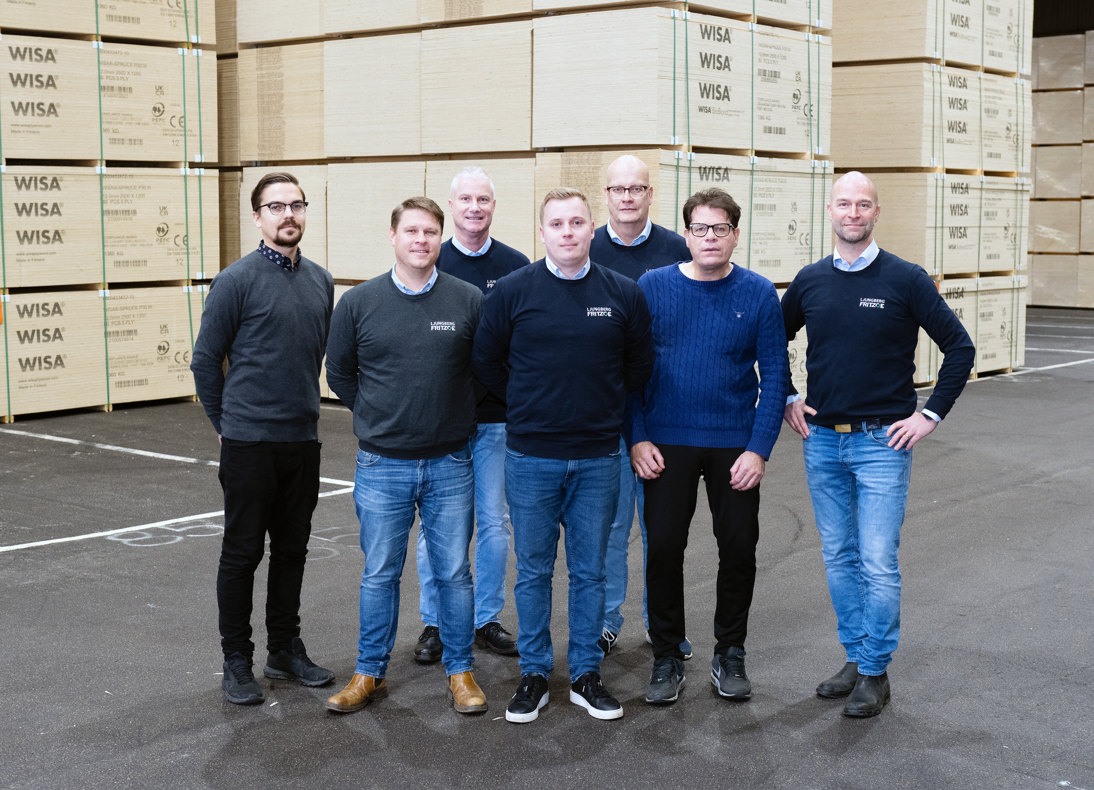 Sju män står tillsammans i en lagerlokal med stora staplar av plywood i bakgrunden. De är klädda i tröjor med företagets logotyp och ser in i kameran.